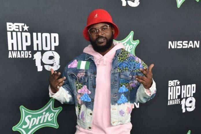 Man wearing a red hat, glasses, and a colorful denim jacket poses with both hands up at the BET Hip Hop Awards 2019 in front of branded event backdrops, channeling the spirit of revolutionary rap and protest music.