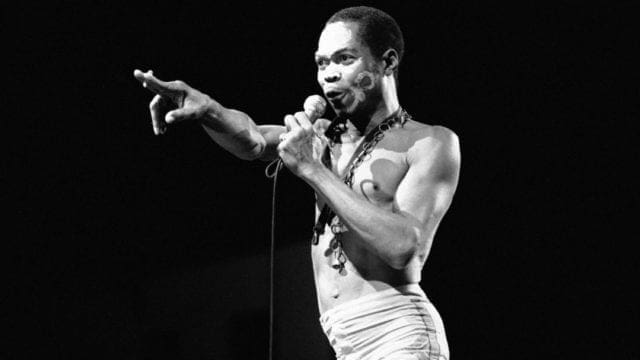 A man holding a microphone performs on stage, wearing minimal clothing and gesturing with his left hand under stage lighting, evoking the energy of Fela, the legendary artist honored with a Grammy Lifetime Achievement Award.