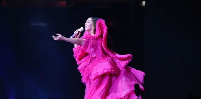 A performer in a bright pink ruffled dress sings into a microphone on stage, channeling Beyoncé’s renaissance era, with one arm extended and dramatic dark background lighting.