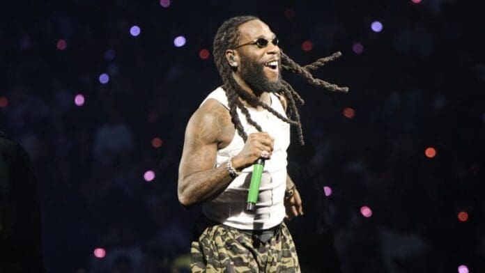 A man with long dreadlocks, sunglasses, and a beard performs on stage, bringing African music into the global spotlight as he holds a green microphone and wears a white tank top and camouflage pants.