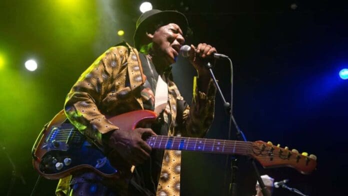 At a festival launch, Ebo Taylor, dressed in a patterned jacket and hat, sings into a microphone while playing an electric guitar on stage under colorful lights—a scene captured by ghanamusic.com.