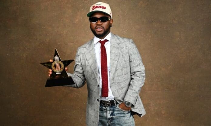 A man wearing a gray plaid blazer, red tie, jeans, sunglasses, and a beige cap holds a star-shaped award—celebrating the 2025 winners at the Telecel Ghana Music Awards—in front of a brown textured background.