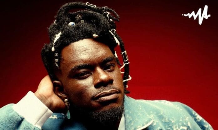 A man with decorated dreadlocks and facial hair poses against a red background in a blue jacket, representing the Up Next Programme for African artists. A white sound wave icon appears in the top right corner, echoing Apple Music’s vibrant energy.