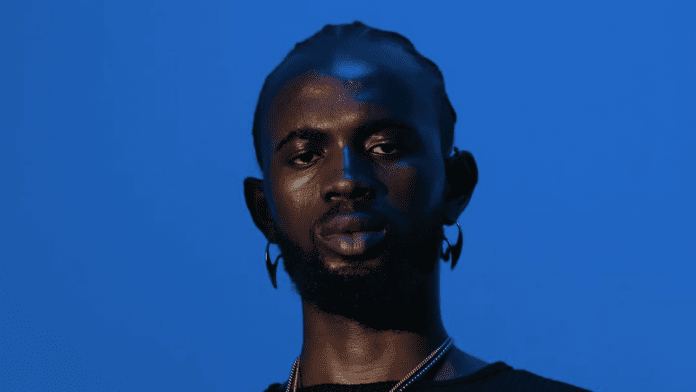 A man with short braids, beard, and hoop earrings wears a black shirt and necklace, channeling Afropop vibes as he poses against a solid blue background—reminiscent of Black Sherif’s Sin City style.
