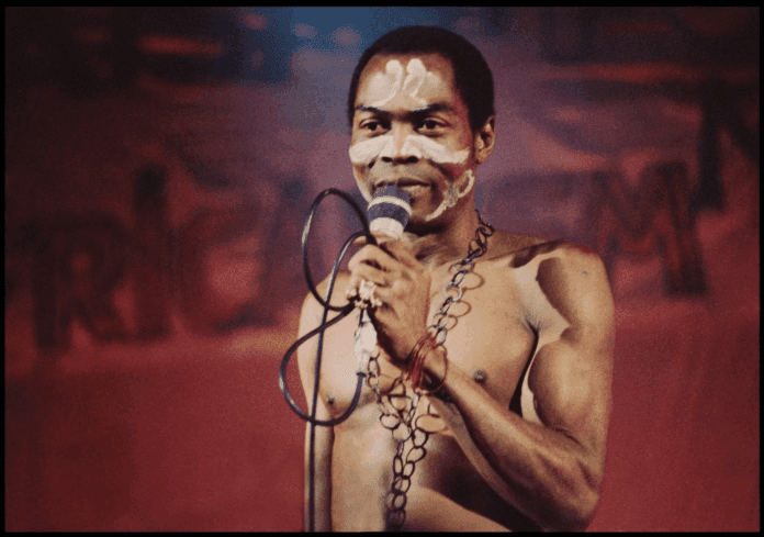 A shirtless man with face paint holds a microphone on stage, wearing a beaded necklace, evoking the bold spirit of Fela, against a red background.