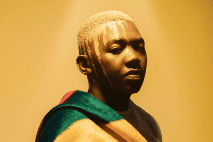 A person with short blond hair and painted white lines on their face stands against a yellow background, draped in a multicolored blanket—an image reminiscent of the vibrant energy at events like the Ngwathe Easter Experience.