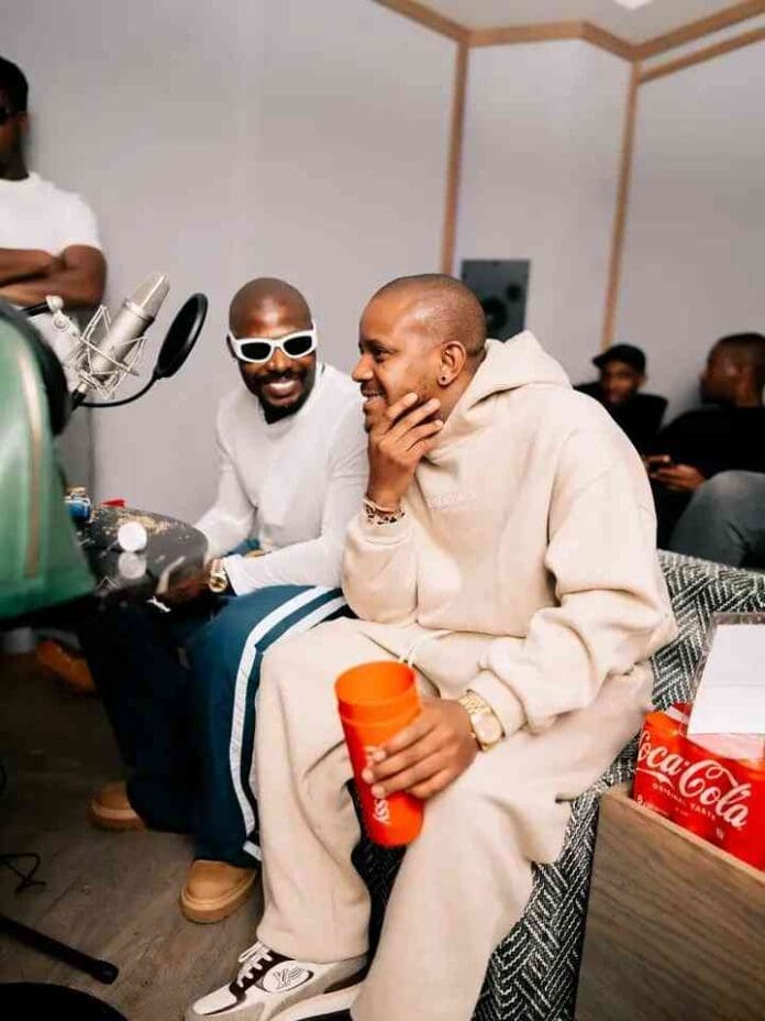 Two men sit and talk in a recording studio; one holds a red cup and smiles, while the other, rocking white sunglasses, looks on. A Coca-Cola box is visible, hinting at a relaxed vibe—perfect for enjoying an Amapiano Mix by Kabza De Small.