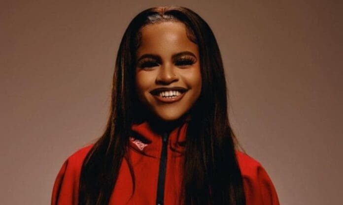 A person with long brown hair smiles at the camera, wearing a red jacket against a plain brown background, capturing the vibrant energy of amapiano—perfect for fans of JayJayy and Phesheya.