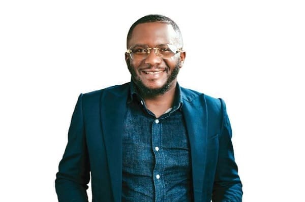 A man in a dark blue blazer and glasses smiles while standing against a plain white background, embodying the confident style often featured in The Nation Newspaper.