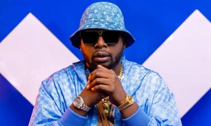 A man wearing a blue patterned bucket hat, sunglasses, and a matching jacket sits with his hands clasped in front of a blue and white geometric background, embodying the fresh style of 2025 inspired by amapiano songs and Music In Africa.