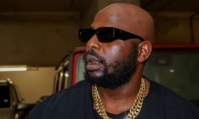 A man with a beard and shaved head wears black sunglasses, a gold chain, gold teeth grills, and a black shirt, standing indoors near a red vehicle at the Milk Closing Festival in Cape Town.