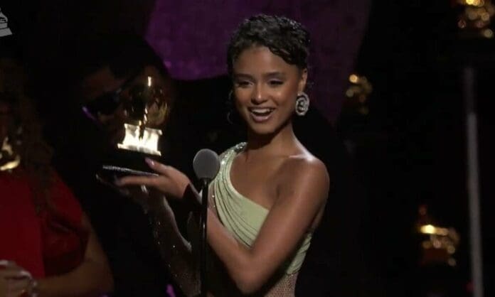 A woman in a light green dress smiles and holds up a gold award trophy while standing at a microphone on stage, celebrating her Grammy Win.