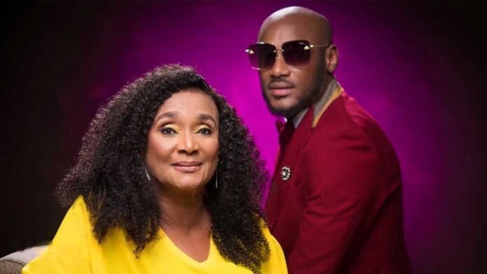 A woman in a yellow top sits in front of a man wearing a red suit and sunglasses, with a purple background behind them, celebrating the greatest Afrobeats songs honoring mothers.