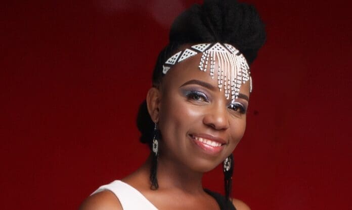 Nokuthula Fundama smiles in a white top against a red background, wearing geometric beaded hair accessories and drop earrings—capturing Joburg’s vibrant spirit after the Heritage concert.
