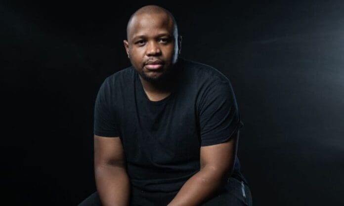 A man wearing a black t-shirt sits against a dark background, looking directly at the camera with a neutral expression, as if ready to share insights from a beginner’s guide to the South African music industry.