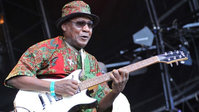 An older man wearing sunglasses and a colorful patterned outfit plays an electric guitar on stage, channeling the vibrant rhythms of Ghanaian highlife in true Ebo Taylor style.