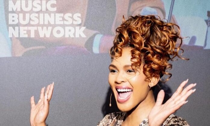 A woman with curly hair smiles and gestures with raised hands while speaking on stage in front of a "Music Business Network" sign, sharing insights about music publishing and industry trends.