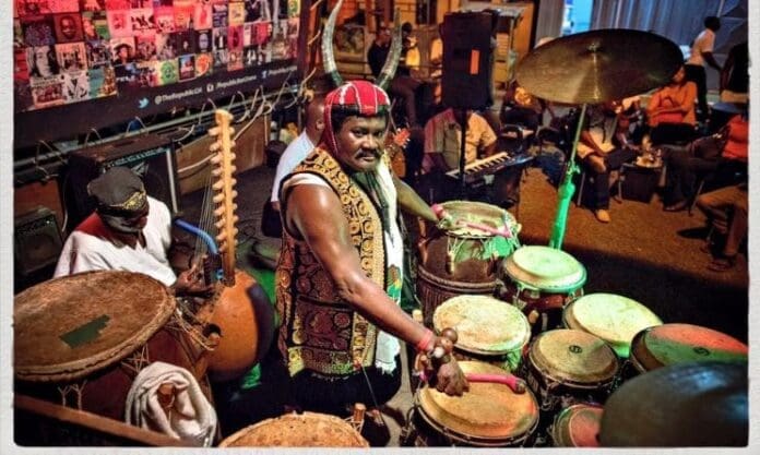 A musician in traditional attire plays a set of drums at an indoor event during Winterjazz 2026, with other musicians and an audience in the background, capturing the vibrant spirit of Music In Africa.