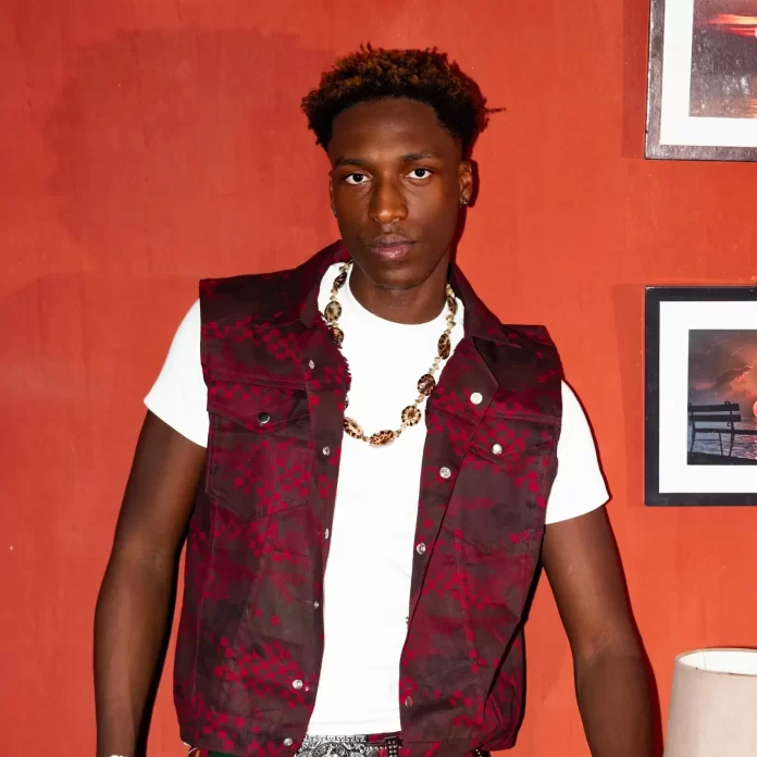 A man in a white t-shirt and patterned maroon vest stands in front of an orange wall with framed pictures, wearing a beaded necklace—a nod to Gen-Z style influenced by Fuji Heritage.
