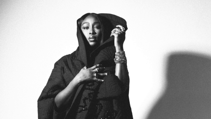 A person in a dark, textured cloak poses with one hand on their chest and the other holding the cloak's hood, against a plain background. The black and white image evokes the soulful artistry found in Tolani’s music, featured on Afrocritik.