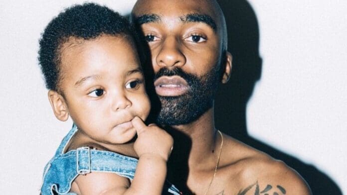 A bearded man holds a young child in denim overalls. Both fathers and children share quiet moments like this, looking slightly off camera as the child rests a finger on their lip.