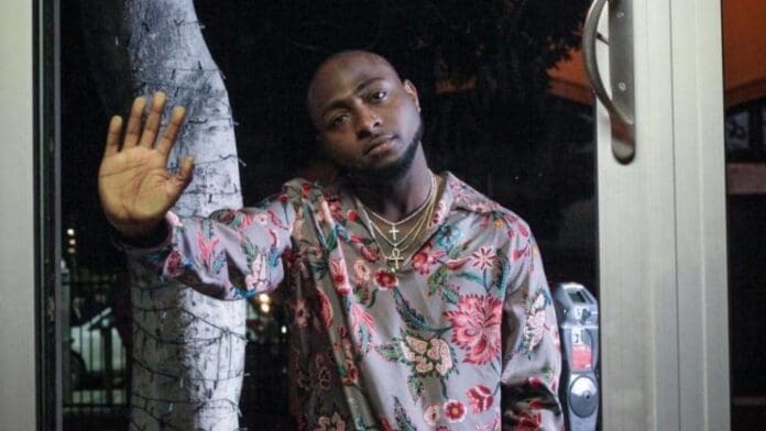 A man in a floral shirt stands behind a glass door at night, raising his hand to the glass and looking out with a neutral expression, as if lost in thought about TV shows or movies he’s seen.