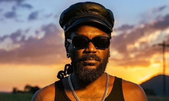 A man wearing sunglasses, a black cap, and a black tank top stands outdoors at sunset, with a mountainous landscape in the background, his earbuds playing Chaana by the Soweto Gospel Choir.