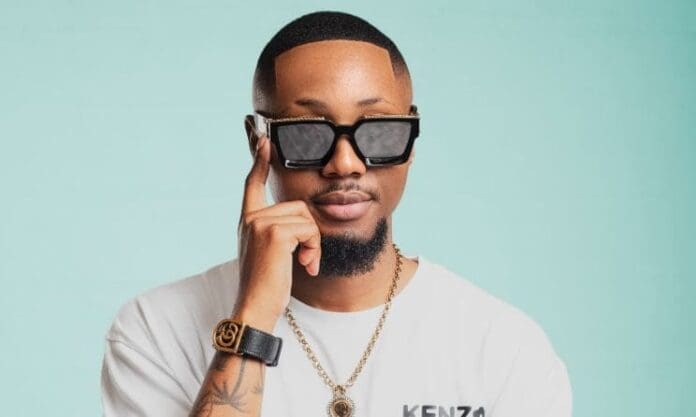 Shaun Stylist, wearing sunglasses, a white Kenzo t-shirt, a gold necklace, and a black wristwatch, poses against a light blue background as he celebrates his second EP praised by Music In Africa.