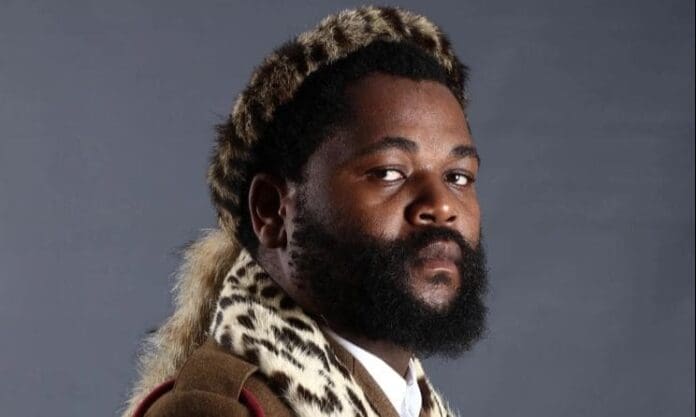 A man with a full beard wears traditional attire with animal print patterns and a matching headdress, channeling the vibrant spirit of Music In Africa as he looks at the camera against a plain gray background.