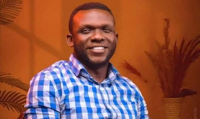 A man wearing a blue and white checkered shirt smiles while seated indoors with plants and an orange background, reflecting the vibrant creativity fueling the African arts scene into a billion-dollar powerhouse.