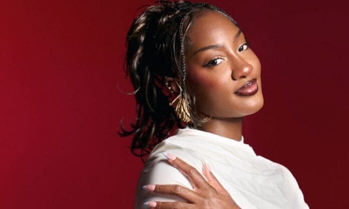 Woman with braided hair and gold earrings poses against a red background, wearing a white top and touching her shoulder—capturing the vibrant spirit of Afrobeats and the colorful energy of Music In Africa.