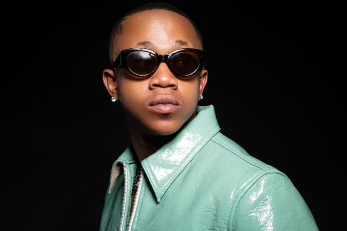 A person wearing dark sunglasses, diamond stud earrings, and a shiny mint green jacket stands against a black background, channeling serious Stush in the Bush vibes.