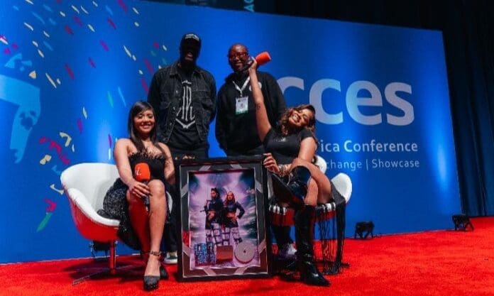 Four people pose on a red carpeted stage at a Music In Africa conference, with TxC seated in front holding microphones, while two others stand behind. A framed photo and awards are proudly displayed before them.