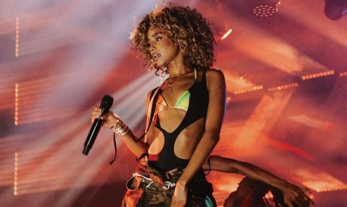 A woman with curly hair holds a microphone and performs on stage, colorful lights and smoke swirling behind her, as part of the 2025 nominees at the Basadi in Music Awards celebrating women in Music In Africa.