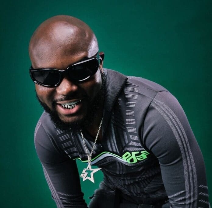 A man with a beard, channeling Afropop vibes, wears dark sunglasses, a gray patterned tracksuit, a star-shaped necklace, and smiles, revealing his grills, against a dark green background.