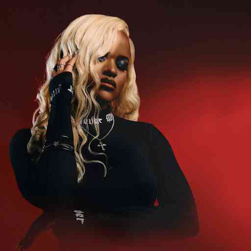 A person with long, wavy blonde hair and dark clothing stands in front of a red background, resting their hand on their face and looking downward, channeling the moody vibes of DJ Tshegu.