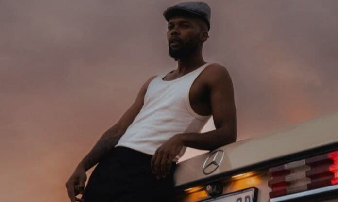 A man in a white tank top and flat cap leans against the back of a beige Mercedes-Benz at sunset, capturing the laid-back vibe of Music In Africa.