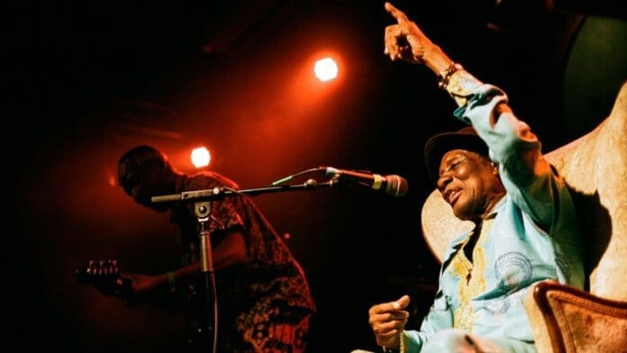 Ebo Taylor, a legend of African highlife music, sings into a microphone on stage, raising one hand, while a guitarist plays beside him under red stage lighting in this NPR performance.
