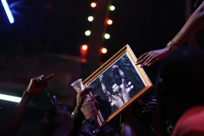 A crowd holds up a framed black-and-white portrait of Fela Kuti during a nighttime event, with colorful lights in the background, celebrating his legacy in African music.