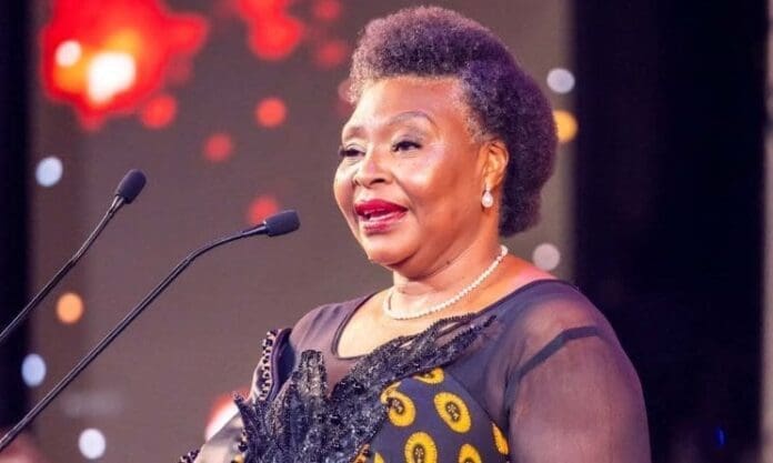 A woman in a patterned dress speaks at a microphone on stage during the Session Music Awards in South Africa, with blurred lights and red accents in the background.