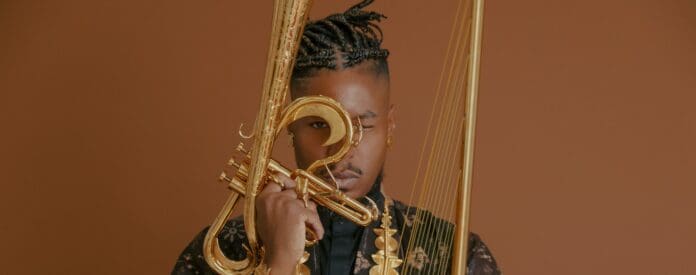 Person holding a golden stringed instrument with a brown background. They have braided hair and are wearing a black outfit with gold accessories, embodying the spirit of Black Indians. The scene evokes the innovative essence of stretch music championed by Chief aTunde Adjuah.