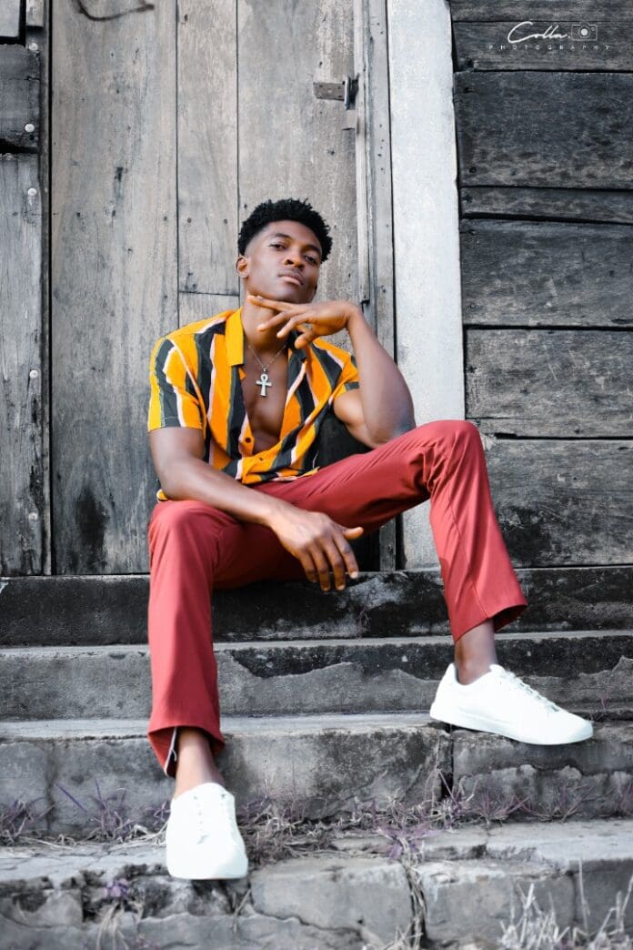 A person sits on stone steps in front of a wooden door, wearing a yellow and black striped shirt, red pants, white sneakers, and a cross necklace. They rest their chin on their hand and look at the camera. Could this be the next Ghana music sensation posing for the Shout-Out EP by K Junior?