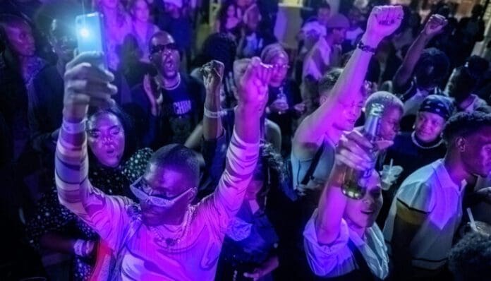 A large group of people in a crowded indoor setting, illuminated by purple lighting, enjoying African hits from 2022, with some raising their arms and holding drinks while one person holds up a phone.