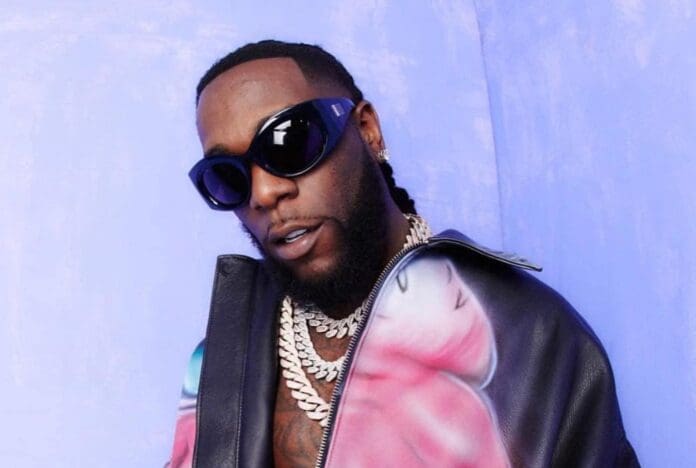 A person with sunglasses and chains, wearing a black leather jacket, stands against a purple background, exuding the same cool vibe as Burna Boy at his Jamaica concert.