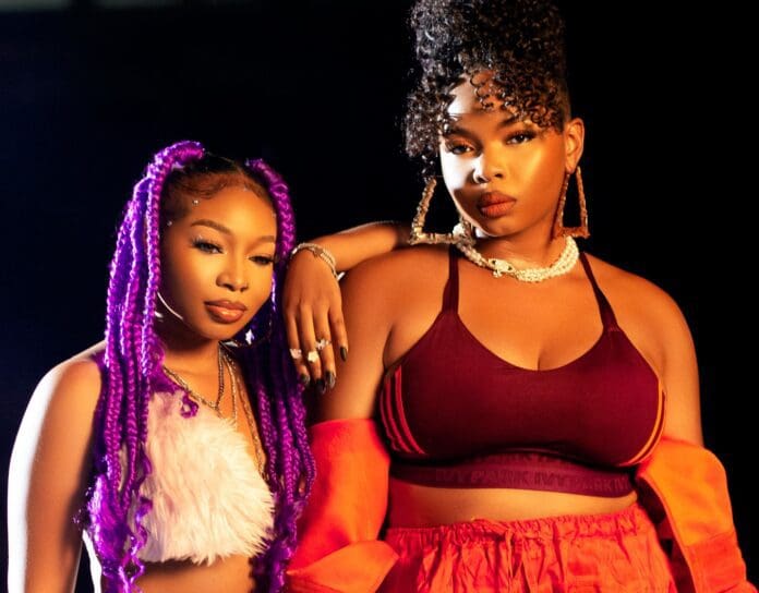 Two women posing confidently, one with long purple braids and a white top, and the other with curly hair, wearing a maroon sports bra and an orange jacket—channeling vibes that could easily be from a Yemi Alade music video.