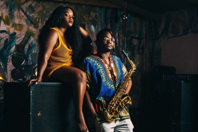 A woman in a yellow dress sits on an amplifier, and a man in a colorful shirt stands next to her holding a saxophone. Both are posing in a dimly lit room with abstract art on the wall behind them, perfectly capturing a Night Time Fall vibe.