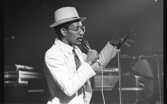 A man wearing a hat and glasses sings into a microphone on stage, gesturing with his hand. He is dressed in a white suit, reminiscent of the iconic style of Linton Kwesi Johnson during one of his memorable five dubs performances.