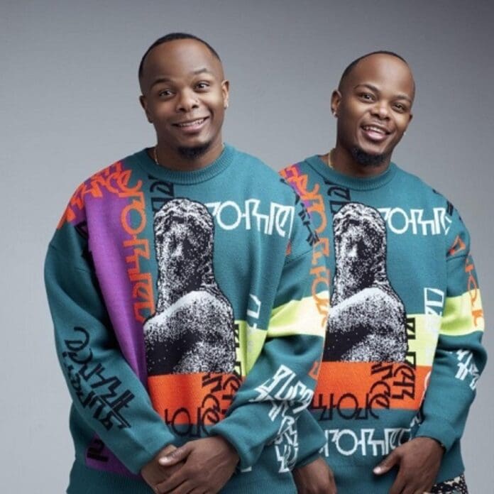 Two men with short hair, side by side, wearing identical colorful sweaters featuring a graphic print of a statue and abstract designs, pose against a plain grey background. They could almost be ready for an Amapiano Mondays set.