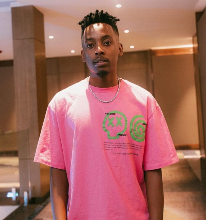 A person stands in a room, wearing a pink T-shirt adorned with artistic designs and text, along with a silver chain necklace that glimmers subtly. The ensemble has an effortless coolness reminiscent of a Daliwonga-inspired vibe.