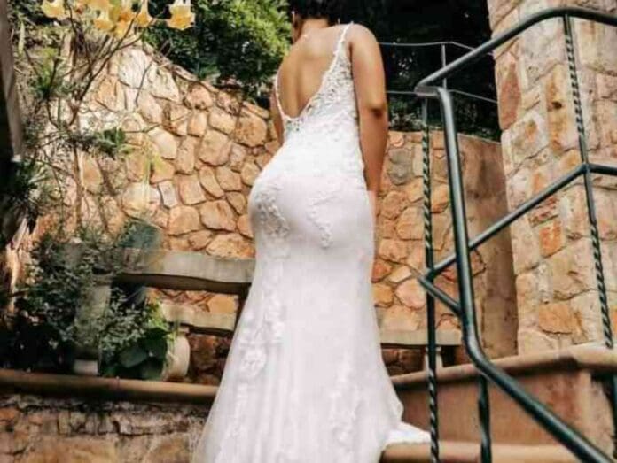 A woman in a white lace wedding dress is seen from behind walking up outdoor stone steps, with greenery in the background, as Sha Sha’s melodious tunes produced by Soa Mattrix elevate the moment.